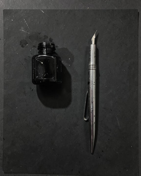 A small black ink bottle sits next to a silver fountain pen on a dark, textured surface.