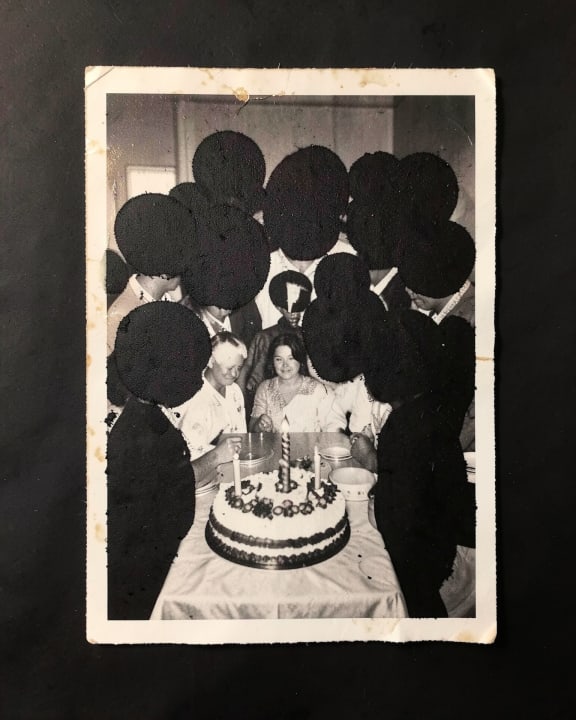 A group of people gather around a birthday cake on a table, though most faces are covered by dark shapes.